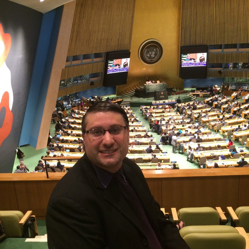 This is a photo of Frank Folino, president of the Canadian Association of the Deaf. The photo is taken at a United Nations meeting. In the background there is a panel of presenters and delegates who are listening.