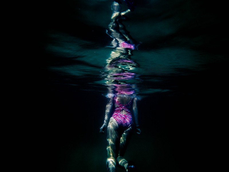 This is a photo of woman in a pink bathing suit submerged under water. Sunlight is reflecting on her body. The image appears to be reflecting above the water but it is distorted and dark. Cest une image dune femme en costume de bain rose dans leau. Le soleil se reflète sur son corps. Limage est nuancée et sombre.